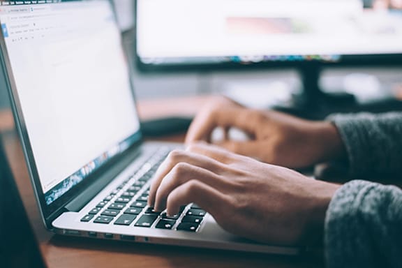 A person is typing on a laptop with a monitor in the background.