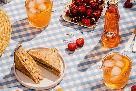 A picnic setup with a sandwich, cherries, and a bottle of aperol spritz.