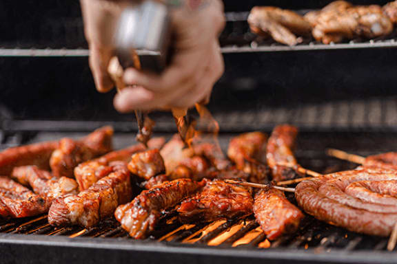 a hand pouring barbecue sauce on chicken wings and sausages on a grill