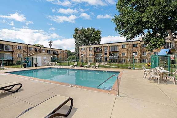 A swimming pool surrounded by chairs and a fence in front of apartment buildings.