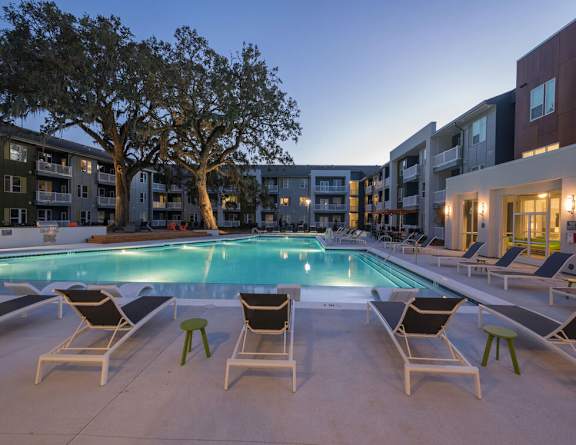 A pool surrounded by chairs and trees with apartment buildings in the background.