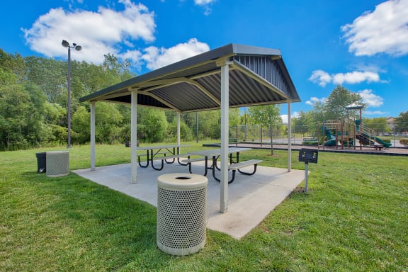 a picnic shelter with a table and benches in a park with a playground in the background