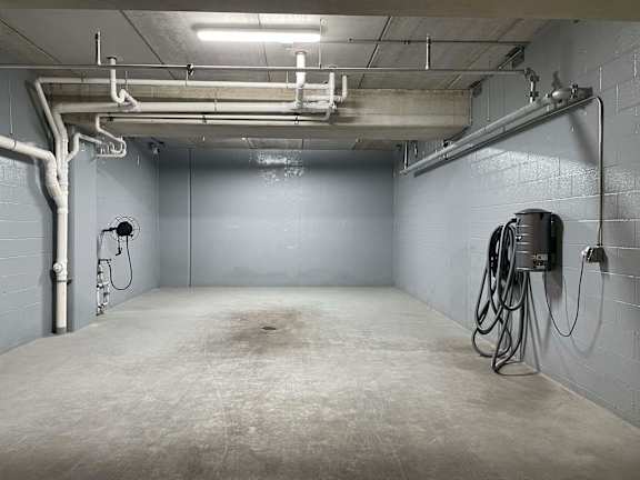 A large, empty room with a concrete floor and walls, equipped with industrial pipes and a concrete mixer.