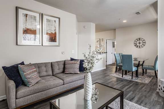 A living room with a grey couch and a white vase on a glass table.