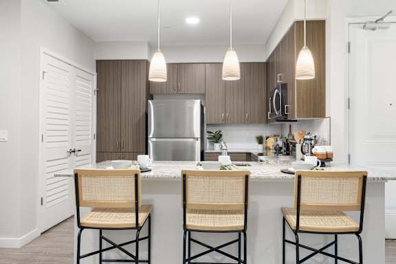 A kitchen with a refrigerator, cabinets, and a counter with three chairs.