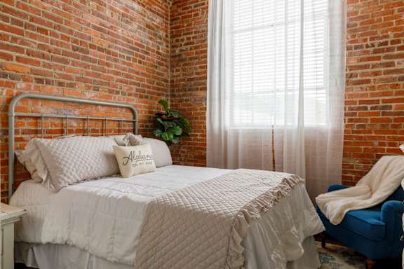 A bed with a white comforter and pillows with the word Alabama on them sits in a room with a brick wall and a window with white curtains.