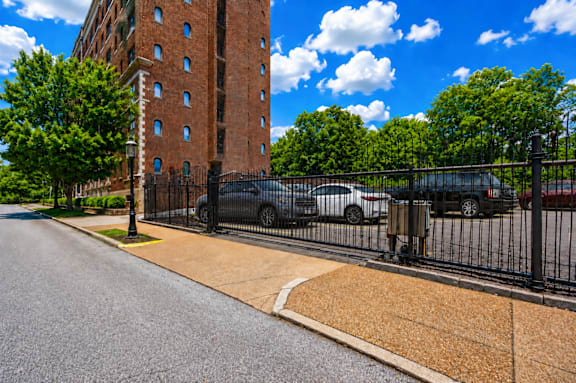 A tall red brick building is behind a black fence and there are cars parked behind the fence.