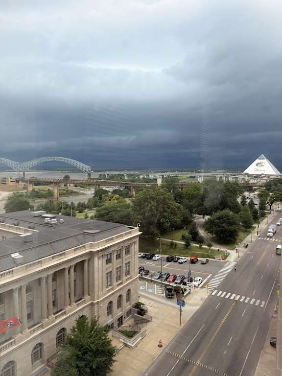 A view of a city street with a large building on the left and a bridge in the distance under a cloudy sky.