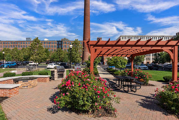 A red brick archway with flowers in front of a parking lot and a building in the background.