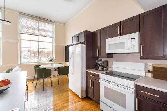 a kitchen with brown cabinets and white appliances and a table with chairs at Beverly Village for Living & the Arts Apartments, Beverly, 01915