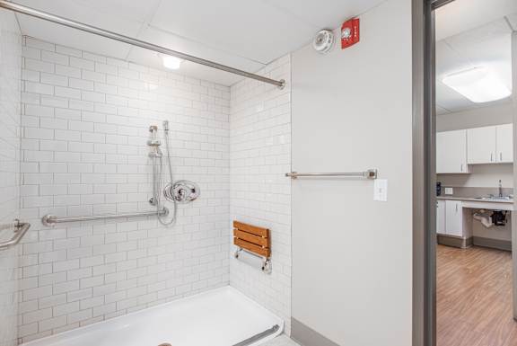 A white tiled bathroom with a walk-in shower and a wooden stool at Moorhead Tower Apartments, Pittsburgh, PA, 15213