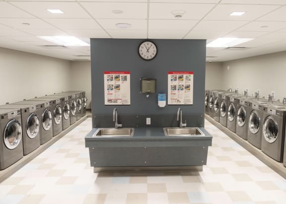 A laundromat with a row of washing machines and a clock on the wall at Highpoint Towers Apartments, Erie, PA