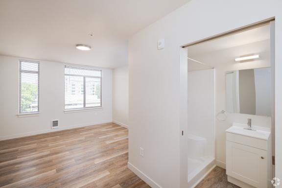 View from entryway of 1-bedroom apartment showing bathroom with a shower and white sink and living room with luxury vinyl floors