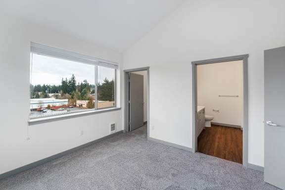 A room with a grey carpet and a view of a parking lot through a window.