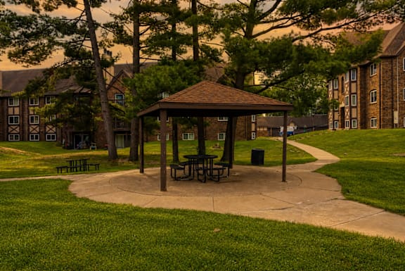 A gazebo with picnic tables is surrounded by trees and grass in a park.