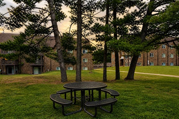 A picnic table sits in the middle of a grassy area with trees and apartment buildings in the background.