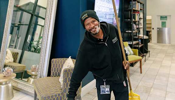 a man in a black hoodie and beanie holds a broom and smiles at the camera