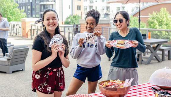 Three people holding food items in front of a picnic table.