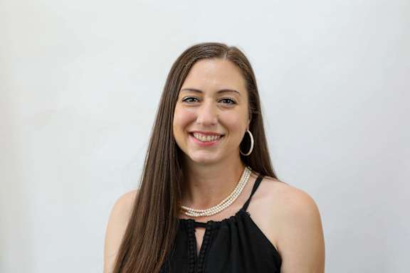 A woman with long brown hair wearing a black top and a pearl necklace.