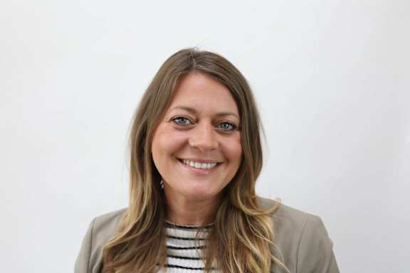 A woman with long brown hair and a striped shirt under a beige blazer.