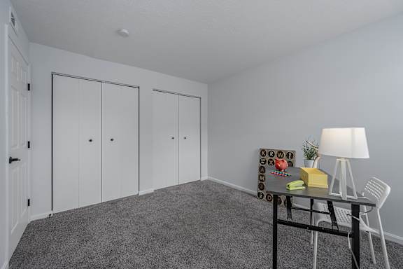 A room with a grey carpet, a white chair, a table with a lamp and a box on it, and three white doors at Indian Lookout Apartments, Ohio, 45449