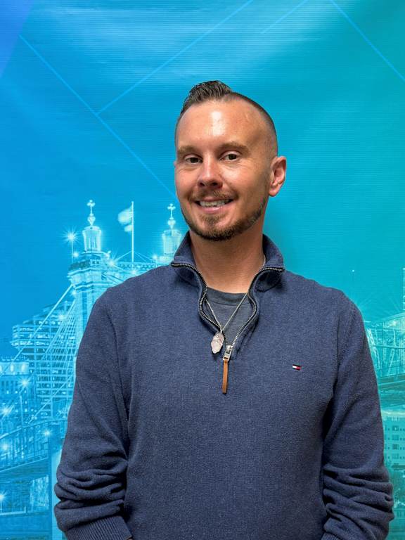 A man with a necklace and a blue shirt is standing in front of a blue background with a building.