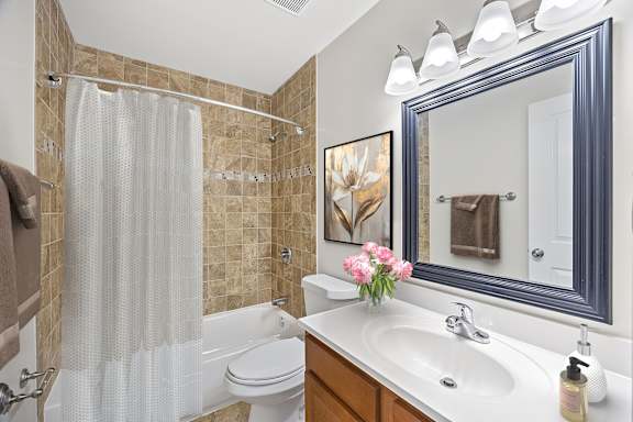 A bathroom with a white sink, a white toilet, a white shower curtain, a brown tile wall, a blue mirror, and a painting of a flower.