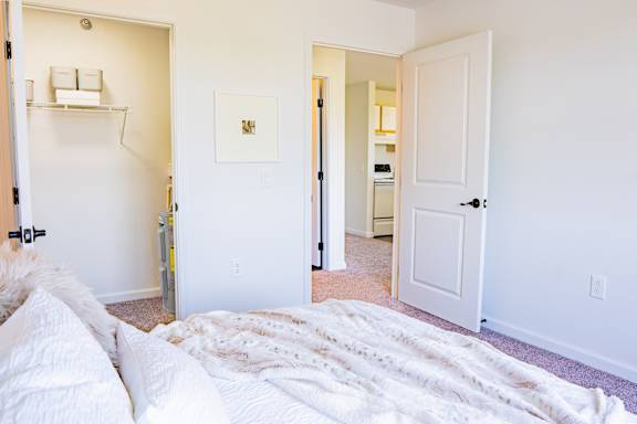 A white bedroom with a bed in the foreground and a hallway leading to a bathroom.