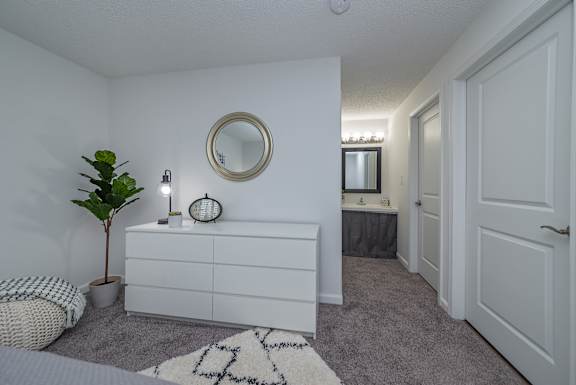 A white bedroom with a round mirror and a plant on the dresser.