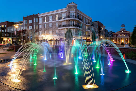 A fountain with multi-colored lights in the foreground with a building in the background.