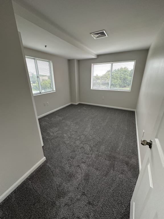 an empty room with gray carpet and two windows at Carisbrooke at Manchester Apartments, New Hampshire