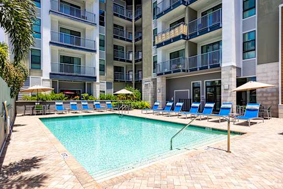 A swimming pool surrounded by blue lounge chairs and umbrellas in front of a multi-story apartment building.