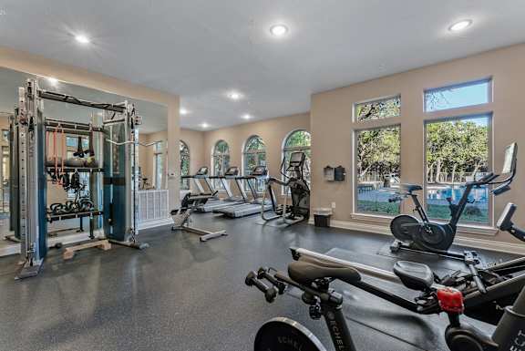 A spacious gym with a variety of exercise equipment and a view of the outdoors through large windows. at Bridge at Heritage Woods, Austin