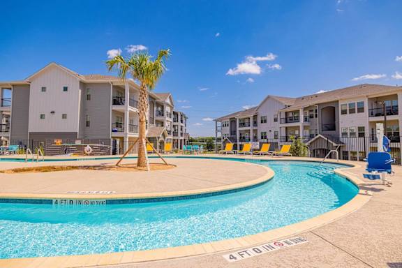 A palm tree stands in the middle of a swimming pool in front of apartment buildings.