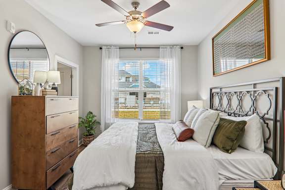 A bedroom with a large bed, a ceiling fan, and a view of a balcony through the window.