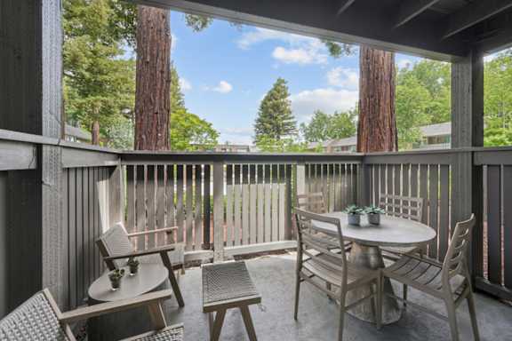 A wooden table with chairs is on a porch with a view of trees.