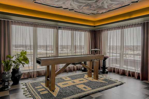 A large wooden table is in the middle of a room with a checkered floor and a large window with curtains.