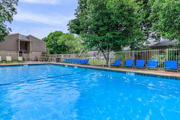 A large blue swimming pool surrounded by trees and a fence with blue lounge chairs.