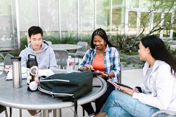 Three students sitting at a table with books and a backpack.