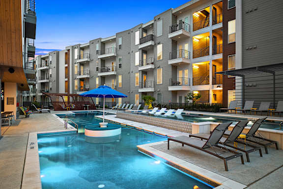 A swimming pool with lounge chairs and umbrellas is surrounded by apartment buildings.