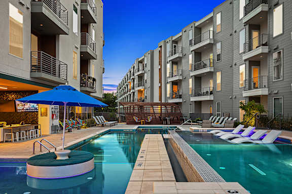A swimming pool with a blue umbrella and lounge chairs in front of apartment buildings.