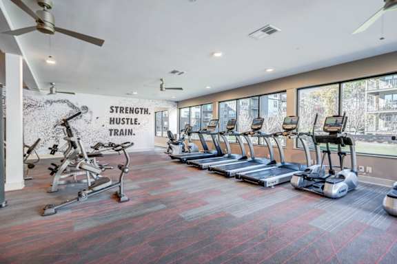 A gym with a variety of equipment including treadmills, elliptical machines, and a wall mural that reads "Strength. Hustle. Train."