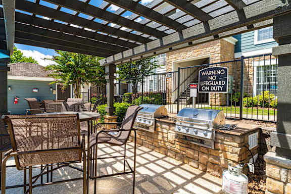 A patio with a grill and chairs under a pergola with a warning sign.