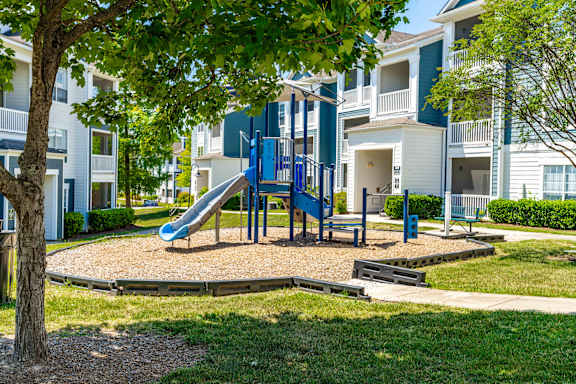 A playground with a blue slide and a green bench in front of apartment buildings.