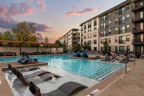 A swimming pool surrounded by lounge chairs and buildings in the background.