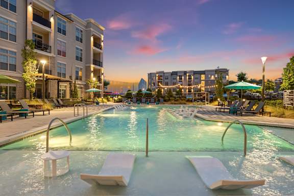 A large outdoor pool with sun loungers and a view of a city skyline at dusk.
