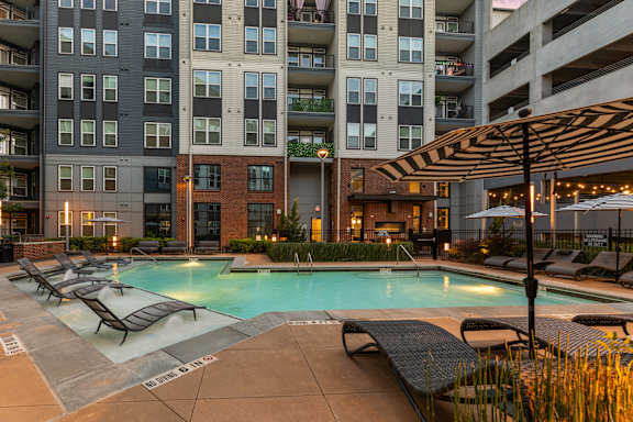 A large swimming pool surrounded by lounge chairs and umbrellas in front of a multi-story apartment building.