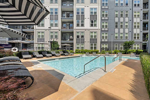 A large swimming pool surrounded by lounge chairs and umbrellas in front of a multi-story apartment building.
