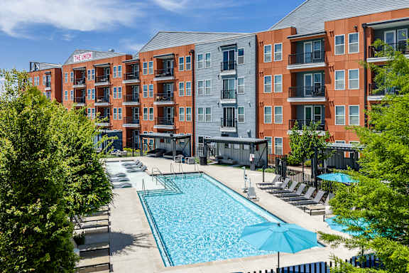 A swimming pool surrounded by lounge chairs and umbrellas in front of a multi-story apartment building.