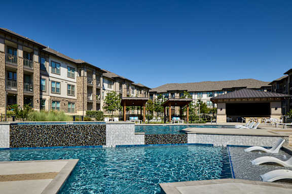 A large swimming pool in front of a building with a stone wall and white lounge chairs.